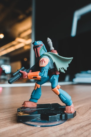 A colorful action figure of a character wearing futuristic armor, equipped with a helmet and a jetpack, is posed on a stand. The character has a blaster in hand and orange accents on the suit, standing out against a blurred background in an indoor setting.