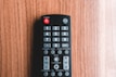 A black remote control with various buttons is placed on a wooden surface. Controls for power, input, numbers, volume, and channel are visible. The wood background has a warm, natural tone.