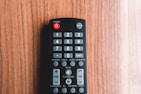 A black remote control with various buttons is placed on a wooden surface. Controls for power, input, numbers, volume, and channel are visible. The wood background has a warm, natural tone.