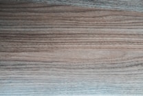 A close-up view of a wooden surface displaying distinct, horizontal grain patterns. The tones vary from light to dark brown, with some subtle grayish hues, giving it a textured and natural appearance.