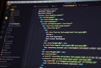 A close-up of a computer screen displaying HTML code in a text editor. The code includes elements such as sections, headers, and paragraphs, with various class attributes for styling. The interface shows file tabs at the top, suggesting it's part of a larger project.