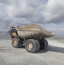 A large industrial dump truck loaded with stones and gravel is positioned in an open, rocky landscape under a cloudy sky. A person in a red outfit is visible near the driver's cabin, providing a sense of scale to the massive vehicle. The setting appears to be a mining site or quarry in a remote area.