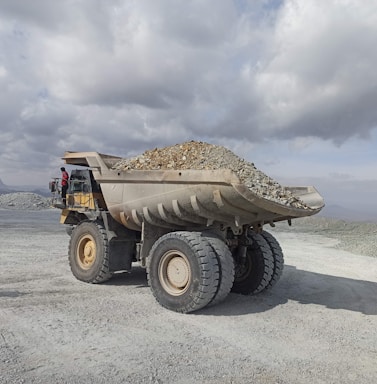 A large industrial dump truck loaded with stones and gravel is positioned in an open, rocky landscape under a cloudy sky. A person in a red outfit is visible near the driver's cabin, providing a sense of scale to the massive vehicle. The setting appears to be a mining site or quarry in a remote area.