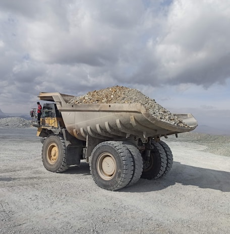 A large industrial dump truck loaded with stones and gravel is positioned in an open, rocky landscape under a cloudy sky. A person in a red outfit is visible near the driver's cabin, providing a sense of scale to the massive vehicle. The setting appears to be a mining site or quarry in a remote area.
