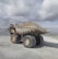 A large industrial dump truck loaded with stones and gravel is positioned in an open, rocky landscape under a cloudy sky. A person in a red outfit is visible near the driver's cabin, providing a sense of scale to the massive vehicle. The setting appears to be a mining site or quarry in a remote area.