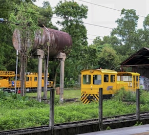A small industrial yellow railway vehicle is positioned near a rusty elevated water tank surrounded by lush green trees and foliage. There is a fence in the foreground and tracks are visible beneath the vehicle. Another similar vehicle can be partially seen in the background near a covered structure.