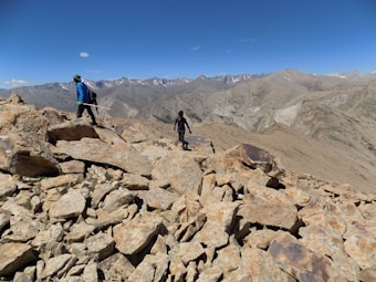 Two hikers traverse a rocky, mountainous terrain under a clear blue sky. The rugged landscape stretches out with rocky outcrops and distant peaks, some with patches of snow. The hikers are equipped with backpacks and walking sticks, indicating a challenging hike or expedition. The expansive view showcases the vastness and raw beauty of the mountainous region.
