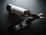 Two wine bottles lying side by side on a dark textured surface. One bottle has a light label with intricate designs and text in a serif font, while the other bottle features a darker label with bold text and a red accent line. Both bottles have corks inserted.