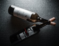Two wine bottles lying side by side on a dark textured surface. One bottle has a light label with intricate designs and text in a serif font, while the other bottle features a darker label with bold text and a red accent line. Both bottles have corks inserted.