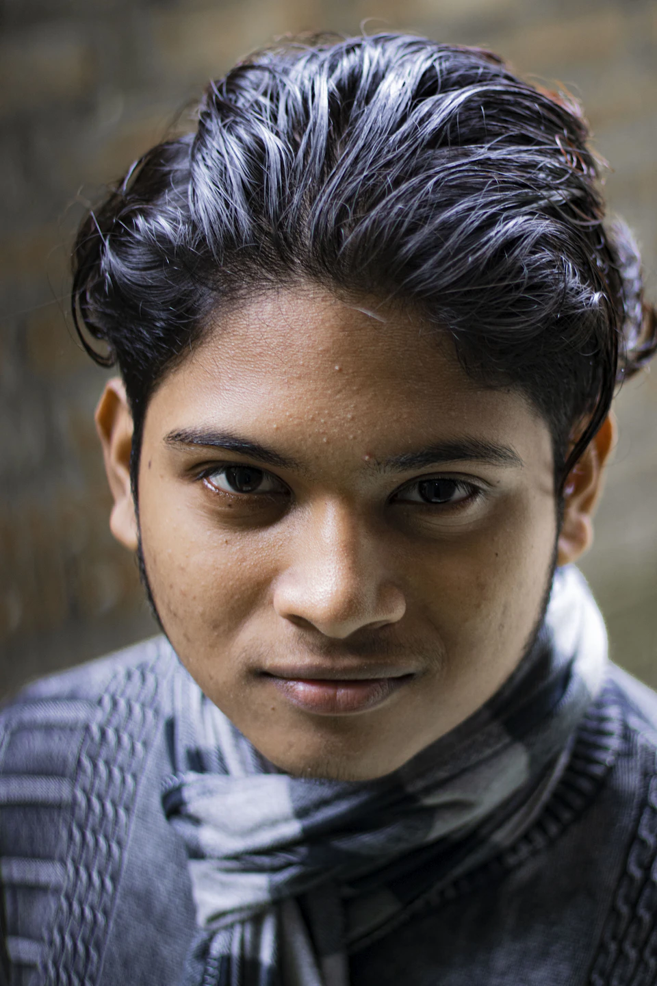 The image shows a young man with dark hair wearing a gray and white striped scarf, looking directly at the camera with an expressionless face. The background is blurred, drawing attention to the subject's features.