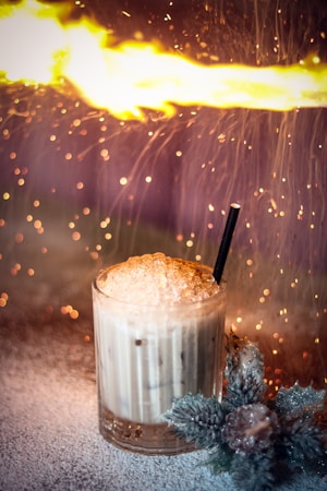 A glass filled with an iced coffee or creamy drink is topped with frothy whipped cream and features a black straw. Surrounding the glass are decorative, frosty, artificial flowers. In the background, a dramatic flame and sparkles create a warm, energetic atmosphere.