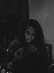 A person is seated indoors, illuminated softly by light, and appears focused on a smartphone. The environment is dimly lit, creating a moody atmosphere. Shadows and highlights emphasize the person's expression and posture.