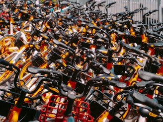 A densely packed collection of rental bicycles, all tightly arranged in rows. The bicycles feature bright colors such as yellow and orange, with noticeable black handlebars and baskets. Each bike is identical in style and design, suggesting they belong to a popular bike-sharing program. The arrangement suggests a high volume of available bikes ready for use.
