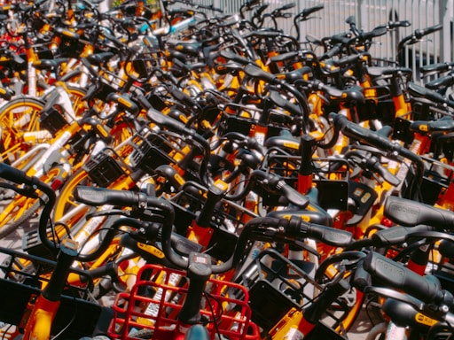 A densely packed collection of rental bicycles, all tightly arranged in rows. The bicycles feature bright colors such as yellow and orange, with noticeable black handlebars and baskets. Each bike is identical in style and design, suggesting they belong to a popular bike-sharing program. The arrangement suggests a high volume of available bikes ready for use.