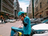 A person wearing a blue uniform and helmet is standing on a busy street talking on a phone. They are leaning on a delivery bike with a turquoise delivery box attached to it. The street is lined with parked cars and tall buildings, and several pedestrians and vehicles are visible in the background.