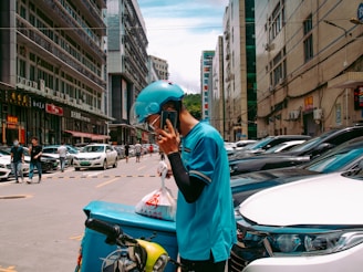 A person wearing a blue uniform and helmet is standing on a busy street talking on a phone. They are leaning on a delivery bike with a turquoise delivery box attached to it. The street is lined with parked cars and tall buildings, and several pedestrians and vehicles are visible in the background.