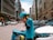 A person wearing a blue uniform and helmet is standing on a busy street talking on a phone. They are leaning on a delivery bike with a turquoise delivery box attached to it. The street is lined with parked cars and tall buildings, and several pedestrians and vehicles are visible in the background.