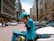 A person wearing a blue uniform and helmet is standing on a busy street talking on a phone. They are leaning on a delivery bike with a turquoise delivery box attached to it. The street is lined with parked cars and tall buildings, and several pedestrians and vehicles are visible in the background.