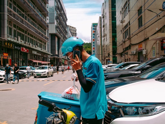 A person wearing a blue uniform and helmet is standing on a busy street talking on a phone. They are leaning on a delivery bike with a turquoise delivery box attached to it. The street is lined with parked cars and tall buildings, and several pedestrians and vehicles are visible in the background.