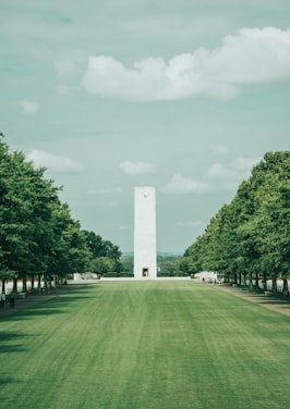 A large, open green lawn extends towards a tall, rectangular white tower. The scene is flanked by rows of lush, green trees under a lightly clouded sky. A few people are seen walking along the pathways on either side of the lawn.