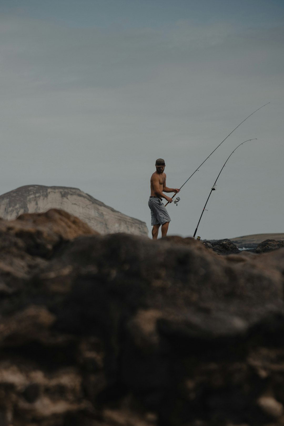 man in white t-shirt and blue denim shorts holding fishing rod standing on brown rock