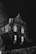 A gothic-style building is illuminated at night, casting intricate shadows from bare tree branches against its facade. The structure features tall, arched windows that are lit from within, creating a stark contrast against the dark night sky.