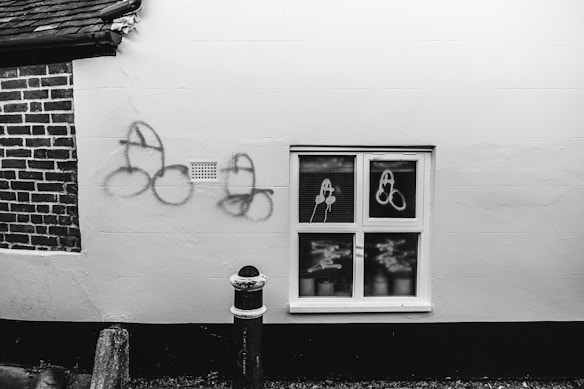 A monochrome image of a building's exterior wall, featuring graffiti on the wall and on a window. The graffiti consists of several drawings with a recurring thematic style. An adjacent window divides into six small panes with similar markings visible. A section of exposed brickwork adds contrast next to the smooth wall.