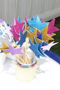 A collection of vibrant, glittery stars attached to wooden sticks is arranged in a cylindrical container. The stars are made of shiny materials and colored in shades of blue, pink, gold, lavender, and silver. They are set on a white surface with a green grass background, partially visible on the side.