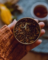A hand in an orange knitted sweater is holding a small tin filled with loose tea leaves and spices. The background features a blurred cup of tea and a hint of yellow, possibly a fruit or decoration, creating a cozy and warm atmosphere.