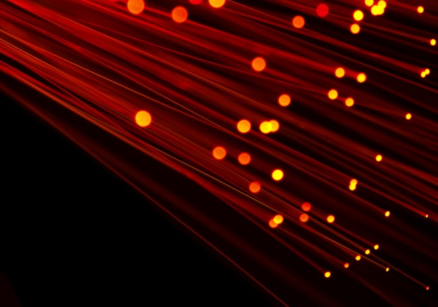 Numerous strands of glowing red-orange fiber optic cables create a dynamic and futuristic visual. The bright points of light accentuate the flowing lines against a dark background, giving a sense of motion.