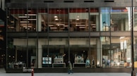 A modern building with large glass windows revealing a bank interior. The exterior reflects city structures, while the ground level showcases posters, screens, and a pedestrian passing by. The bank's logo is visible at the top.