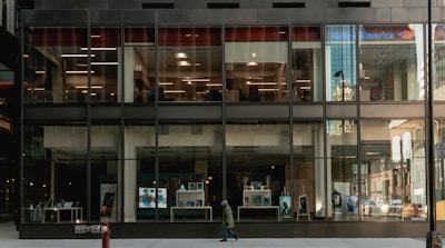 A modern building with large glass windows revealing a bank interior. The exterior reflects city structures, while the ground level showcases posters, screens, and a pedestrian passing by. The bank's logo is visible at the top.