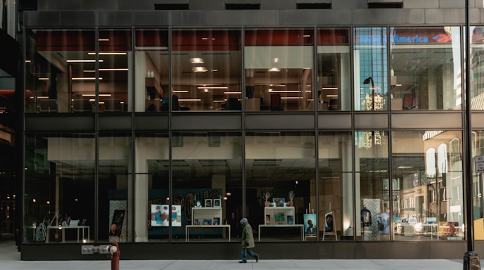 A modern building with large glass windows revealing a bank interior. The exterior reflects city structures, while the ground level showcases posters, screens, and a pedestrian passing by. The bank's logo is visible at the top.
