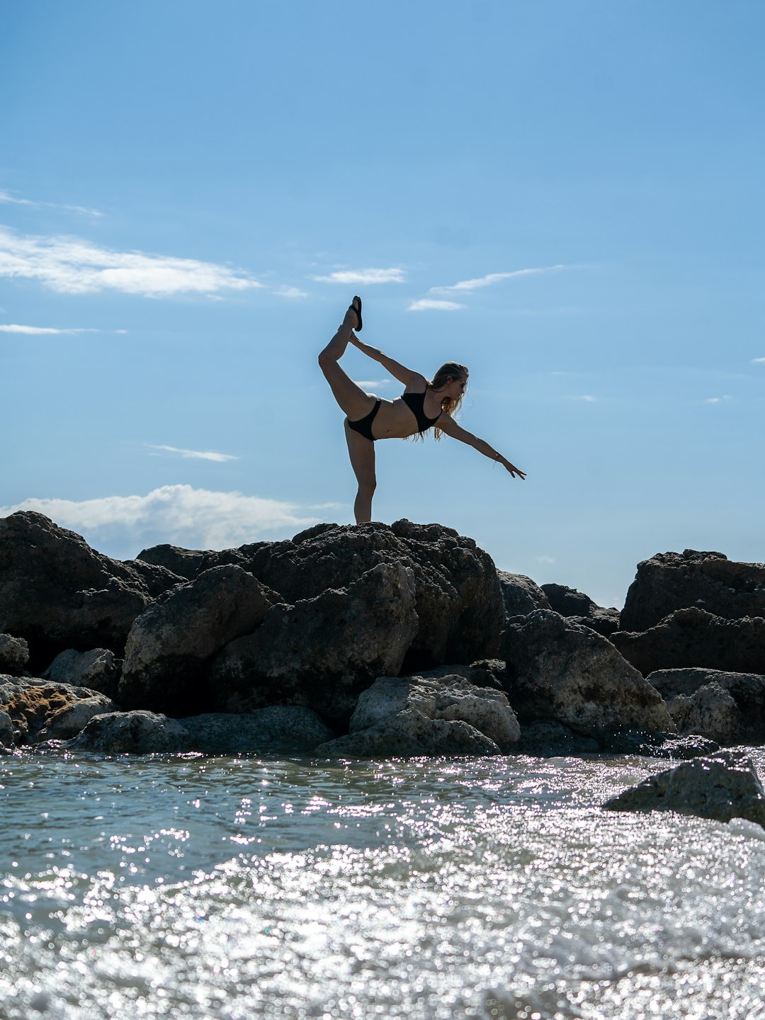 woman in black bikini jumping on rocky shore during daytime