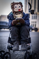A child is sitting in a stroller, wearing a hat with a cartoon monkey face on it. The child is engrossed in a smartphone, holding it with both hands. The child is dressed warmly in a blue jacket, grey pants, and winter boots. The background is an urban setting with blurred buildings and vehicles.
