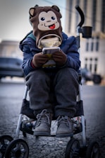 A child is sitting in a stroller, wearing a hat with a cartoon monkey face on it. The child is engrossed in a smartphone, holding it with both hands. The child is dressed warmly in a blue jacket, grey pants, and winter boots. The background is an urban setting with blurred buildings and vehicles.