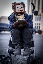 A child is sitting in a stroller, wearing a hat with a cartoon monkey face on it. The child is engrossed in a smartphone, holding it with both hands. The child is dressed warmly in a blue jacket, grey pants, and winter boots. The background is an urban setting with blurred buildings and vehicles.