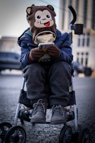 A child is sitting in a stroller, wearing a hat with a cartoon monkey face on it. The child is engrossed in a smartphone, holding it with both hands. The child is dressed warmly in a blue jacket, grey pants, and winter boots. The background is an urban setting with blurred buildings and vehicles.