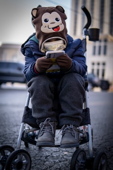 A child is sitting in a stroller, wearing a hat with a cartoon monkey face on it. The child is engrossed in a smartphone, holding it with both hands. The child is dressed warmly in a blue jacket, grey pants, and winter boots. The background is an urban setting with blurred buildings and vehicles.