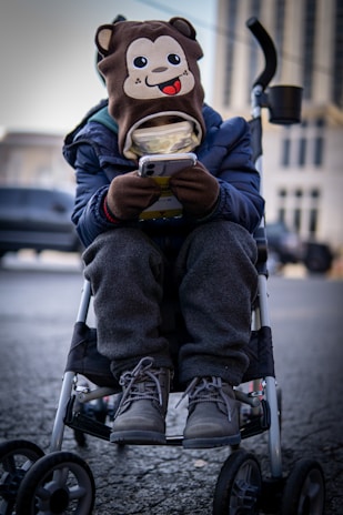 A child is sitting in a stroller, wearing a hat with a cartoon monkey face on it. The child is engrossed in a smartphone, holding it with both hands. The child is dressed warmly in a blue jacket, grey pants, and winter boots. The background is an urban setting with blurred buildings and vehicles.