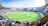A large, open cricket stadium filled with a crowd. The lush green playing field is surrounded by stands packed with spectators. Bright sunlight illuminates the stadium, casting shadows on the ground. Tall floodlights tower above the stands, and advertisements are visible around the stadium's perimeter.