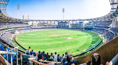 A large, open cricket stadium filled with a crowd. The lush green playing field is surrounded by stands packed with spectators. Bright sunlight illuminates the stadium, casting shadows on the ground. Tall floodlights tower above the stands, and advertisements are visible around the stadium's perimeter.