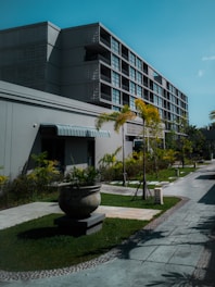 A modern multi-story building with a minimalist design featuring large windows and a textured facade. The foreground includes a well-maintained garden with several potted plants and small palm trees lining a paved walkway. The sky is a clear blue, enhancing the serene and organized environment.