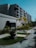 A modern multi-story building with a minimalist design featuring large windows and a textured facade. The foreground includes a well-maintained garden with several potted plants and small palm trees lining a paved walkway. The sky is a clear blue, enhancing the serene and organized environment.