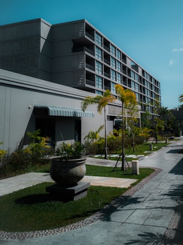 A modern multi-story building with a minimalist design featuring large windows and a textured facade. The foreground includes a well-maintained garden with several potted plants and small palm trees lining a paved walkway. The sky is a clear blue, enhancing the serene and organized environment.