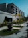 A modern multi-story building with a minimalist design featuring large windows and a textured facade. The foreground includes a well-maintained garden with several potted plants and small palm trees lining a paved walkway. The sky is a clear blue, enhancing the serene and organized environment.