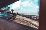 A beachfront resort area featuring a multi-story building on the left side with balconies and palm trees. A pathway runs alongside the shoreline, which is dotted with outdoor seating and loungers. The atmosphere is enhanced by a panoramic view of the ocean under a partly cloudy sky.
