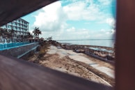 A beachfront resort area featuring a multi-story building on the left side with balconies and palm trees. A pathway runs alongside the shoreline, which is dotted with outdoor seating and loungers. The atmosphere is enhanced by a panoramic view of the ocean under a partly cloudy sky.