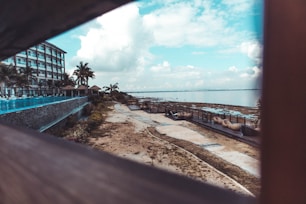 A beachfront resort area featuring a multi-story building on the left side with balconies and palm trees. A pathway runs alongside the shoreline, which is dotted with outdoor seating and loungers. The atmosphere is enhanced by a panoramic view of the ocean under a partly cloudy sky.