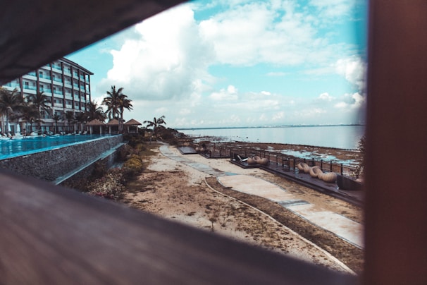 A beachfront resort area featuring a multi-story building on the left side with balconies and palm trees. A pathway runs alongside the shoreline, which is dotted with outdoor seating and loungers. The atmosphere is enhanced by a panoramic view of the ocean under a partly cloudy sky.